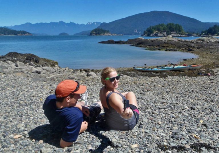 Man and a woman sitting on a rocky beach with kayaks on the shore. Several islands and mountains in the background.