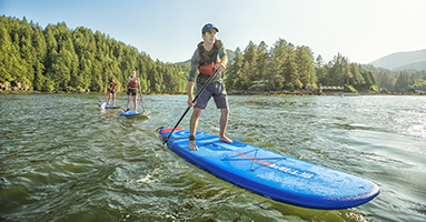 Man on blue stand up paddle board with wooded shoreline and other stand up paddle boarders in the background