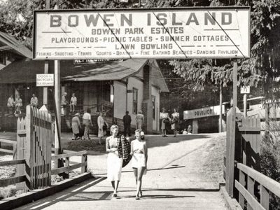 Black and white picture of two women walking on a pier with a pavilion in the background. There is a sign above welcoming them to Bowen Island - Park Estates circa 1948